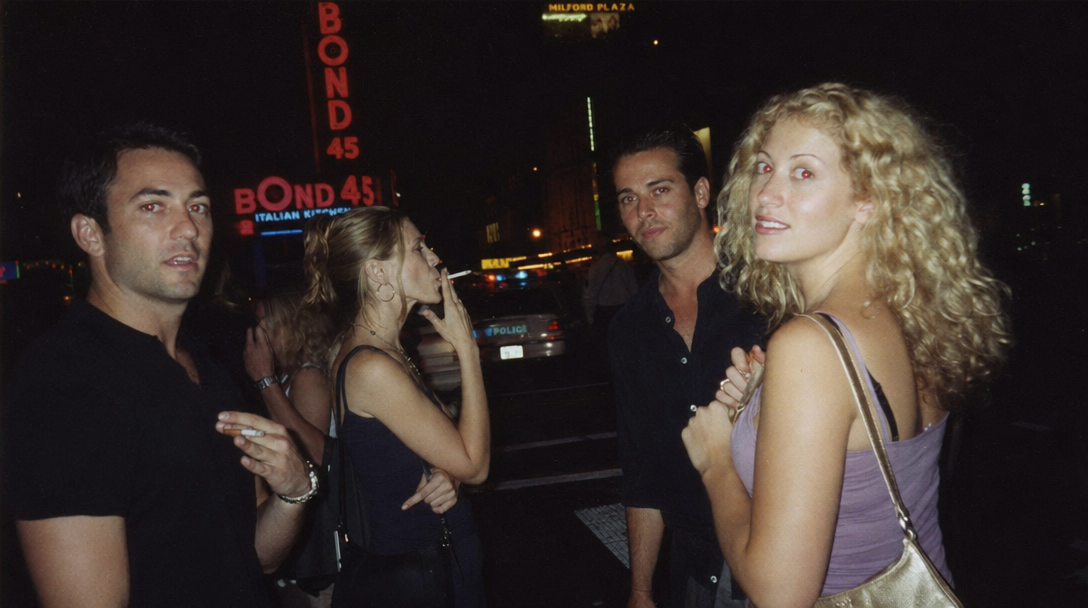 Late-90s tabloid paparazzi shot outside the Bond Bar — group of friends laughing under neon signage, blown-out flash, motion blur, scanned-from-magazine grain, authentic 1999 nightlife mood.