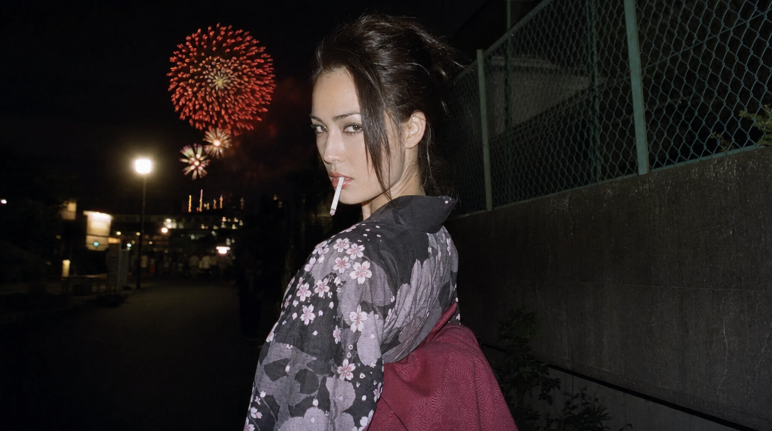 Candid disposable-camera portrait — young woman in floral kimono looking back over her shoulder on a quiet Japanese street, distant hanabi fireworks bursting over rooftops, warm summer-festival nostalgia.